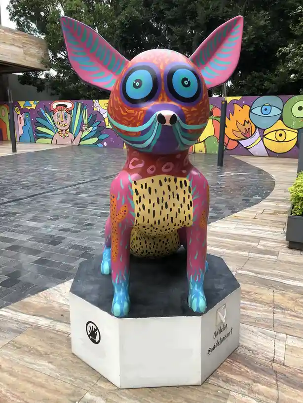 A brightly painted, stylized dog sculpture with oversized pink ears, large blue eyes, and colorful patterns sits on a…