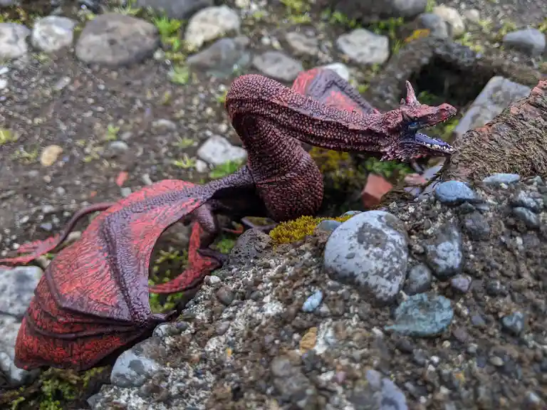 A red, textured plastic dragon toy with wings and horns is positioned on a rocky, mossy ground, appearing to be outdoors…