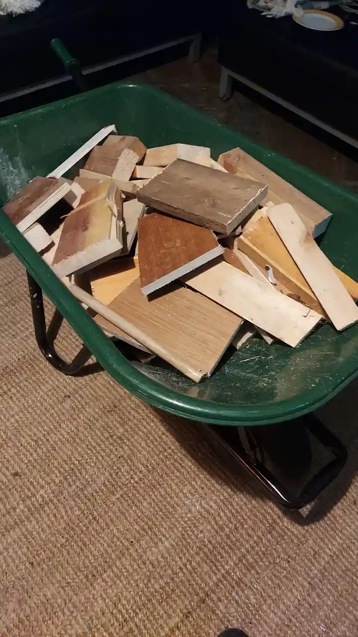 A green wheelbarrow sits indoors on a beige carpet, filled with various pieces of cut wood, including flooring samples and…
