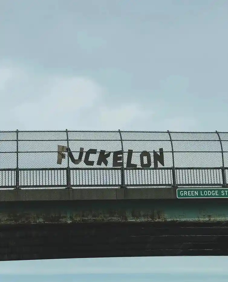 A low-angle photo of a bridge with a green street sign reading 'GREEN LODGE ST' on its side. On the chain-link fence atop…