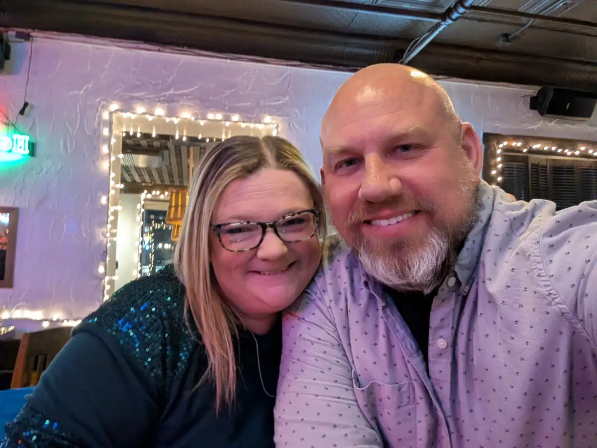 A selfie of a smiling bald man with a beard and a woman with glasses, both looking at the camera in a dimly lit bar with…