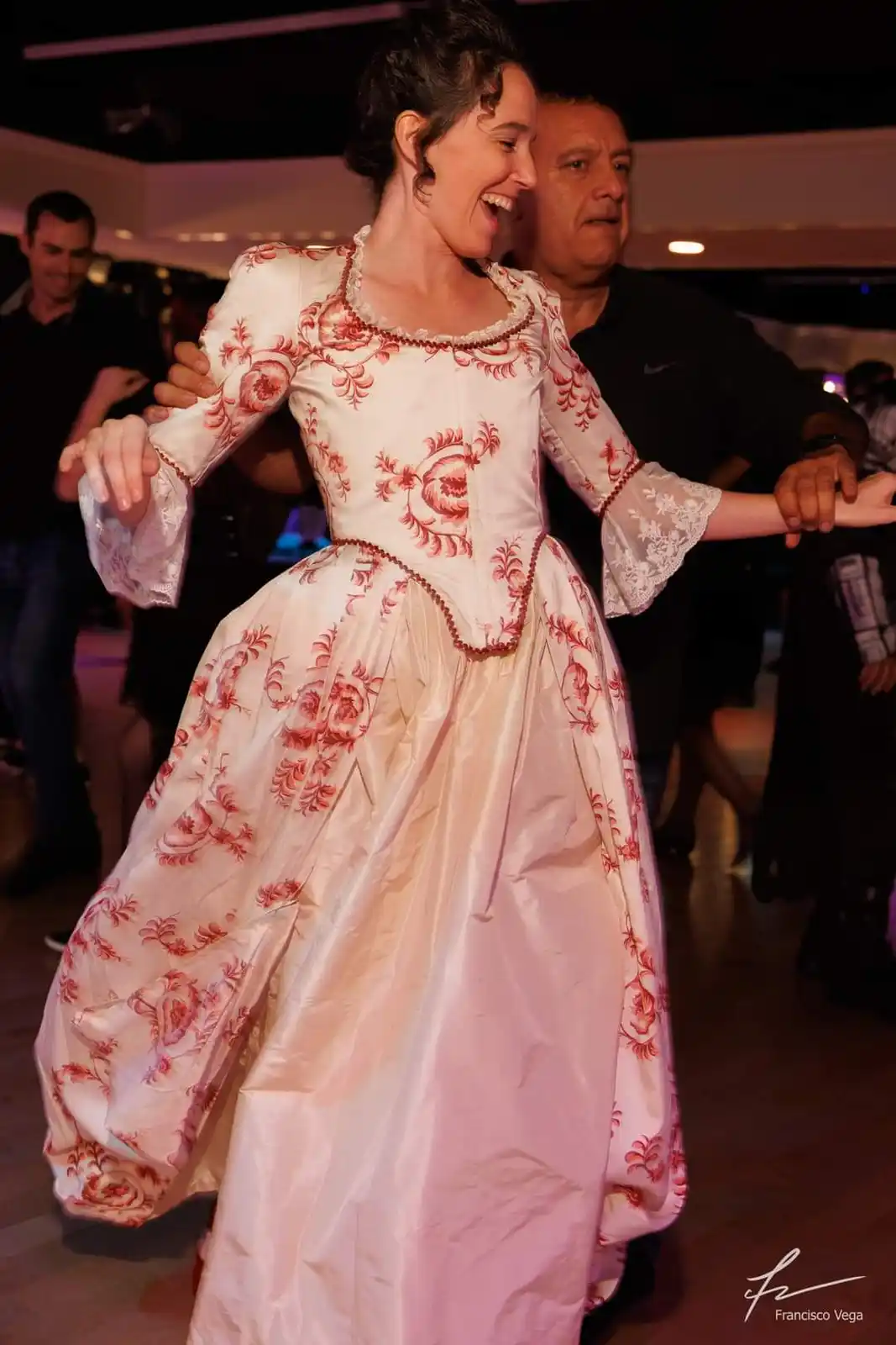 A woman in a white and red floral historical gown is dancing joyfully with a man in a black shirt, both holding hands. The…