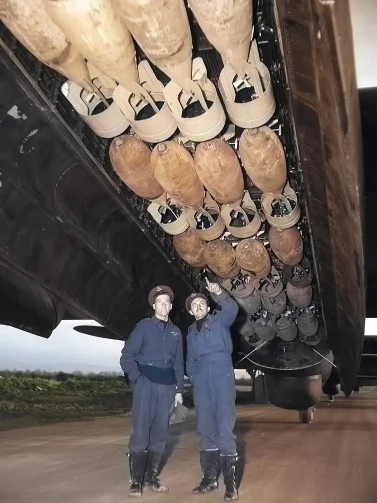 Two men in military uniforms stand beneath the bomb bay of a large aircraft, surrounded by numerous large bombs hanging in…