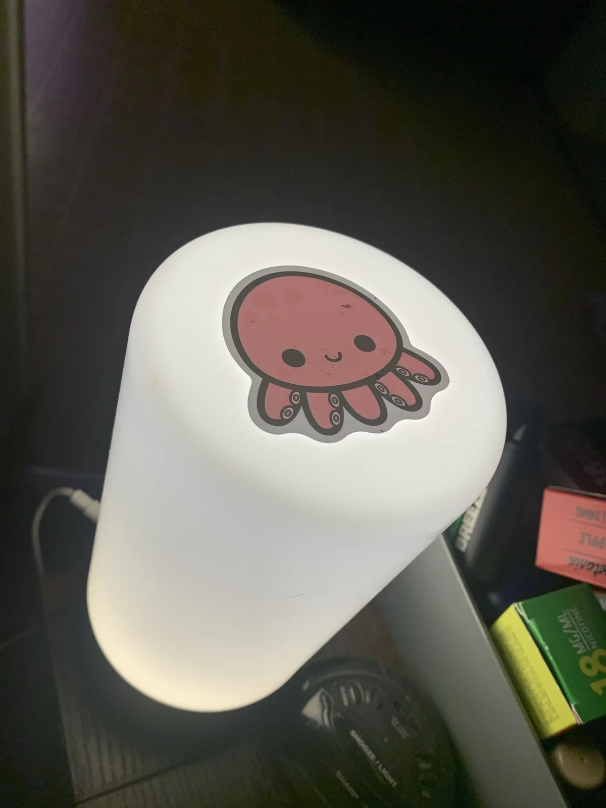 A white, glowing desk lamp with a cute pink cartoon octopus sticker on its top surface, placed on a dark surface with some…