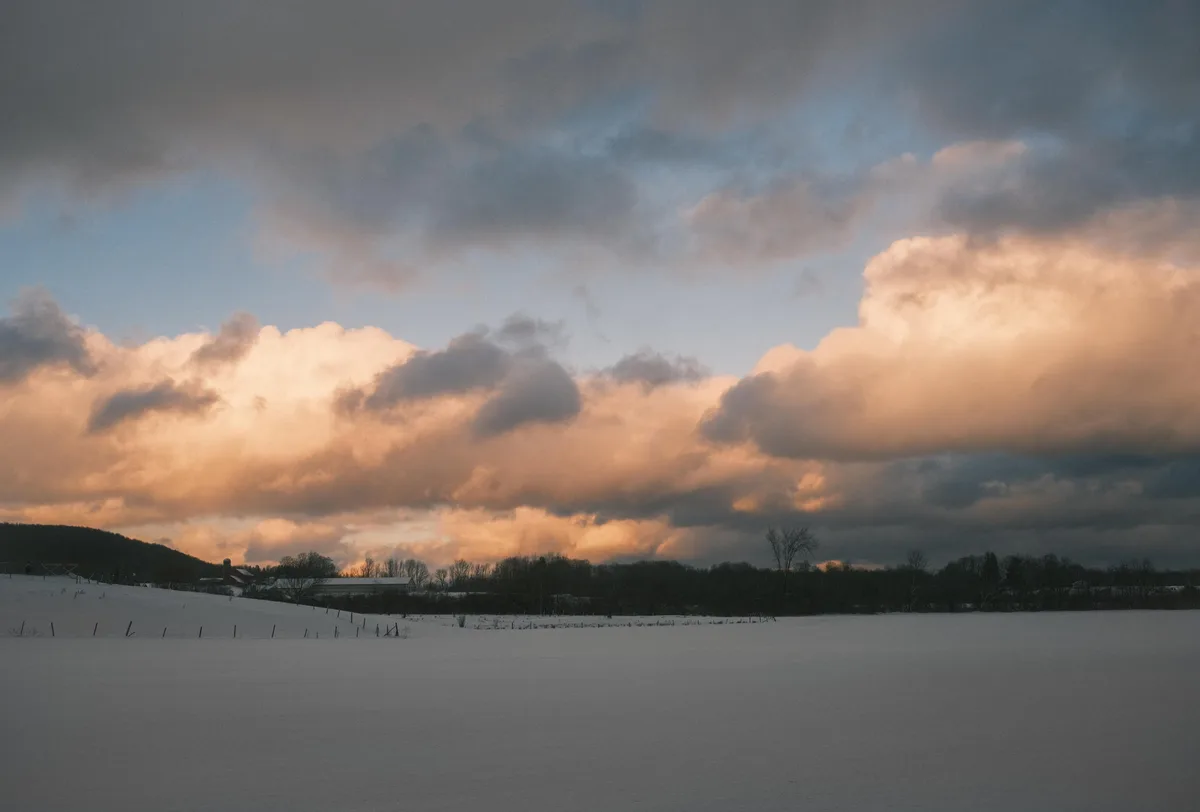 A serene winter landscape shows a vast snow-covered field under a dramatic sky with clouds lit in warm orange and gray tones…
