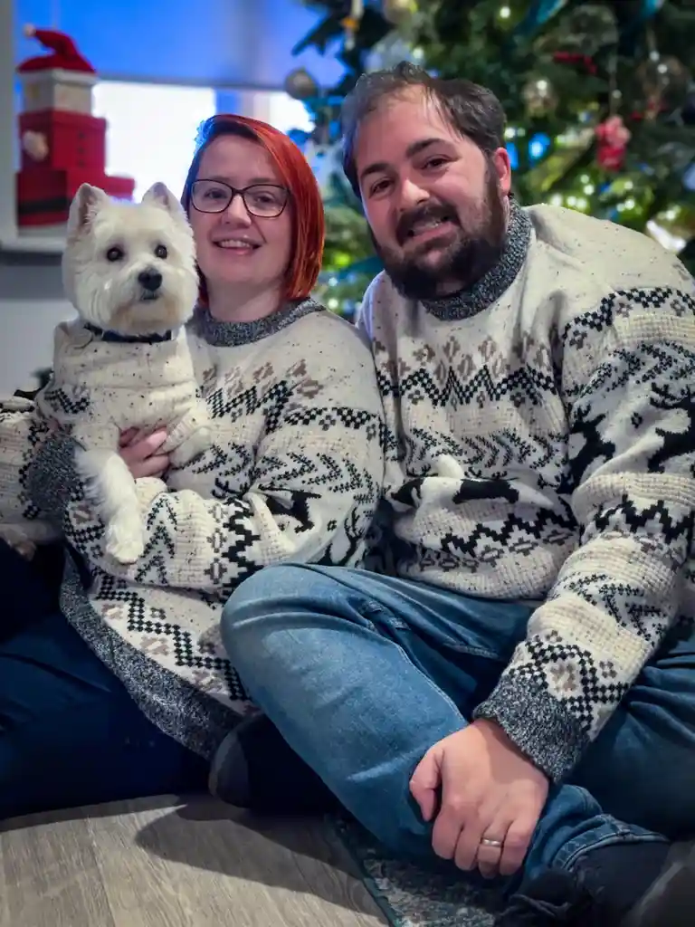A man and woman in matching festive sweaters sit on the floor in front of a decorated christmas tree, the woman holding a…