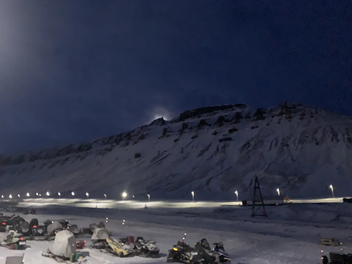 A nighttime photo shows a snowy mountain landscape with a lit-up road and parked snowmobiles in the foreground. The sky is…