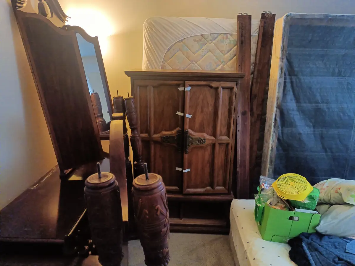 A cluttered bedroom shows a wooden armoire with duct tape holding its doors shut, a bed frame leaning against the wall, and…