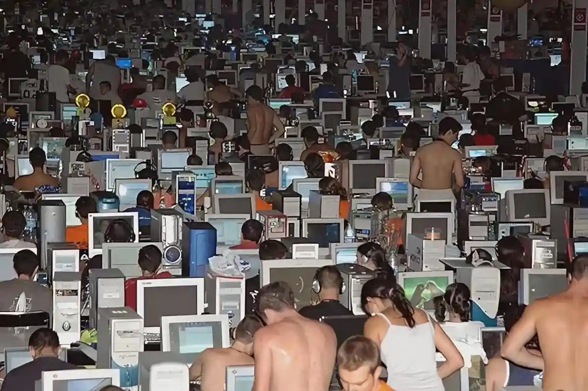 A crowded, dimly lit internet cafe filled with dozens of people sitting at computer stations, many shirtless or in casual…