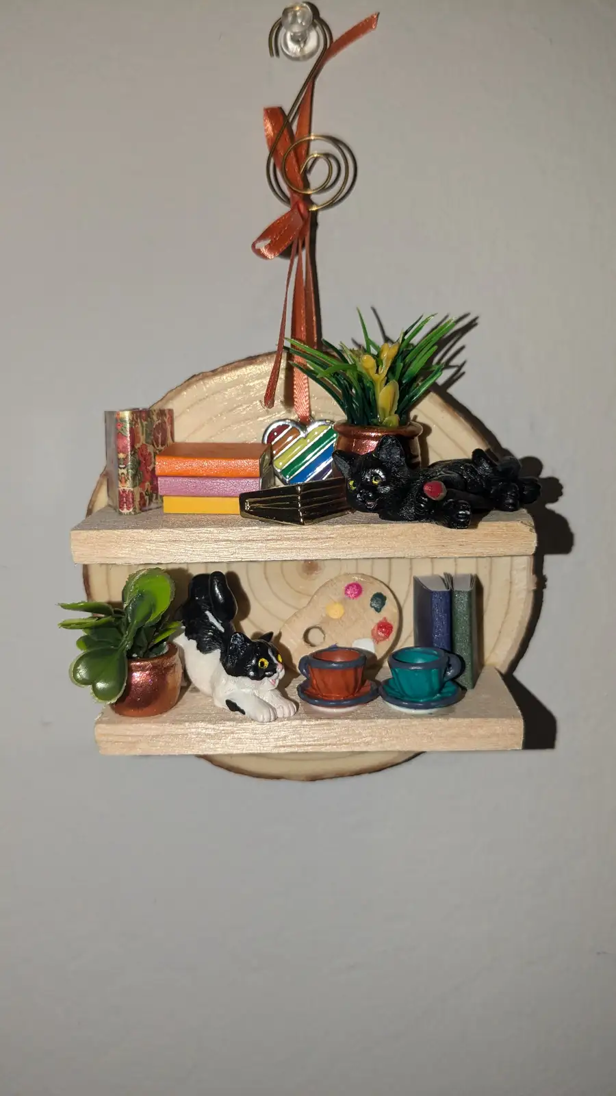 A miniature two-tiered wooden shelf ornament with a black cat figurine lounging on the top shelf beside books and a potted…