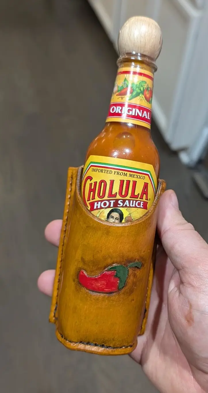 A hand holds a bottle of Cholula Original Hot Sauce, which is encased in a brown leather holster designed to look like a gun…