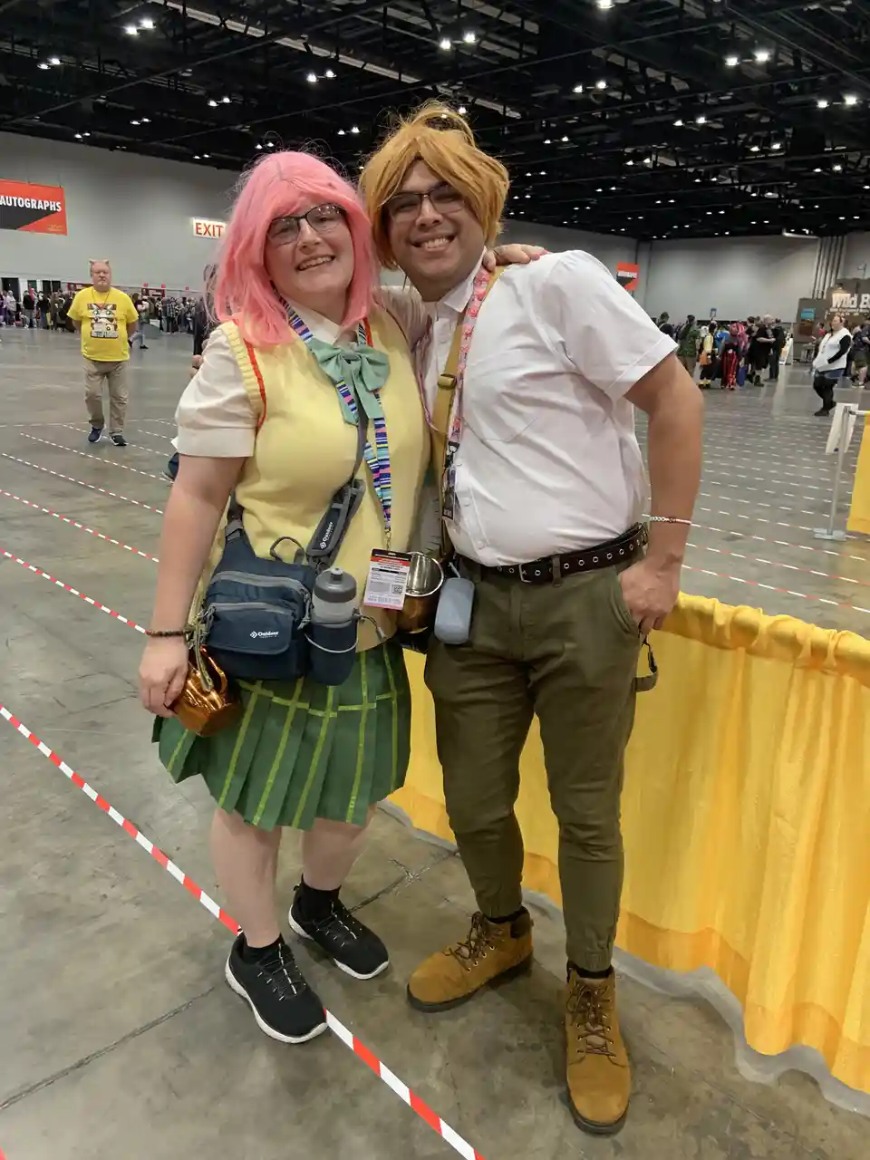 Two people in cosplay pose together at a convention; one wears a pink wig, glasses, a yellow vest, green pleated skirt, and…