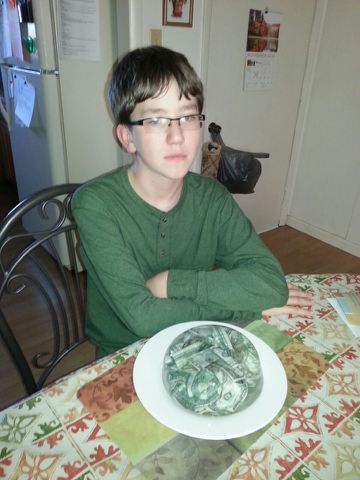 A young person with glasses sits at a dining table with arms crossed, looking unimpressed. In front of them is a white plate…