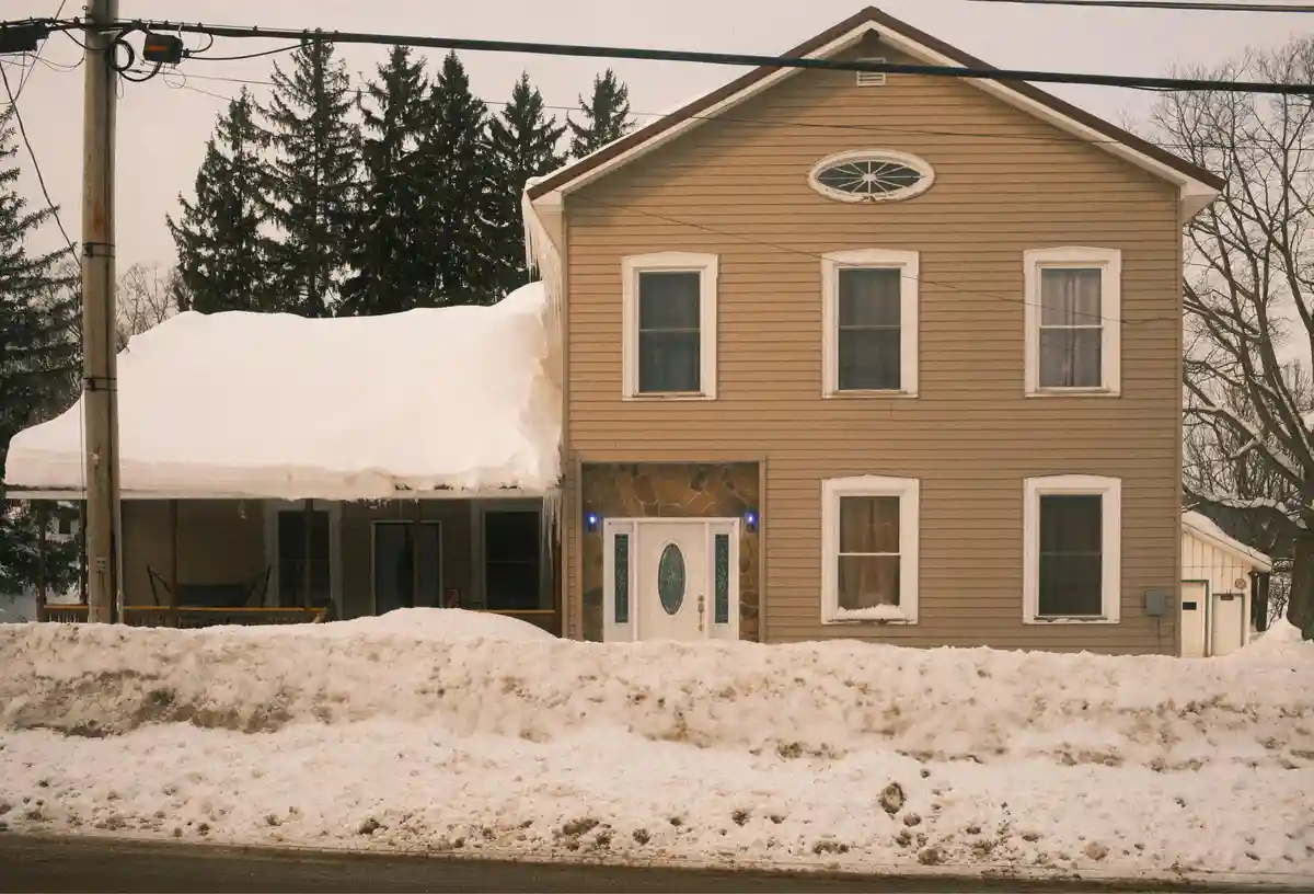 A two-story beige house with snow piled high on its porch roof and along the front yard, with icicles hanging from the…