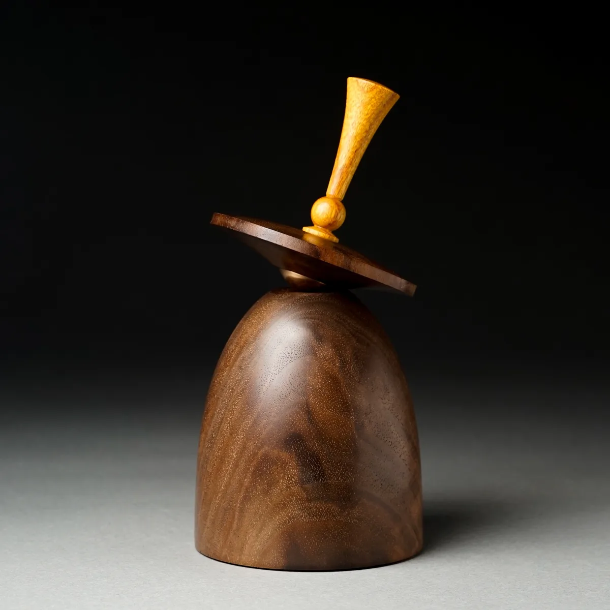 A wooden sculpture resembling a minimalist hourglass or bell with a handle-like top, set against a dark background. The…