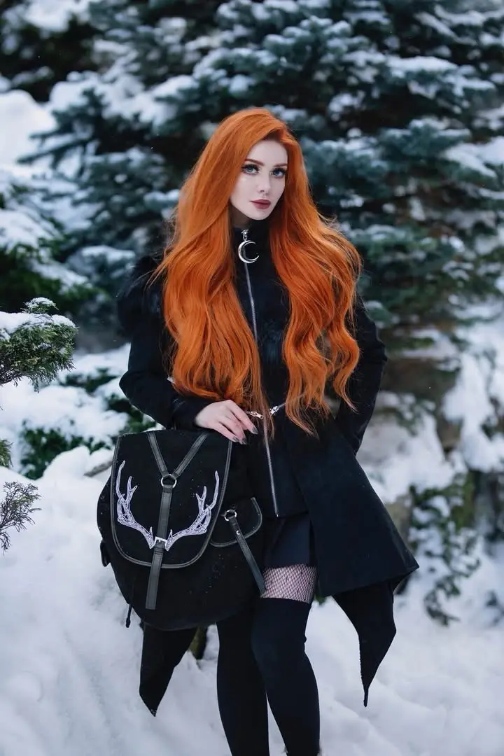 a woman with long, vibrant orange hair stands in a snowy forest wearing a black coat, fishnet stockings, and a black…