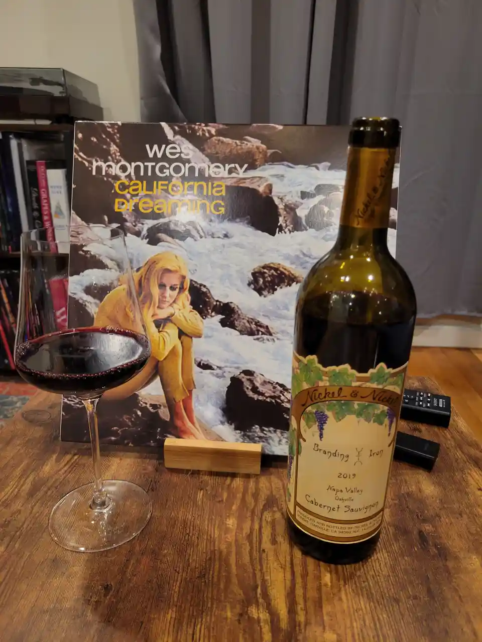 A bottle of Nickel & Nickel 2019 Cabernet Sauvignon and a glass of red wine sit on a wooden table in front of a vinyl record…
