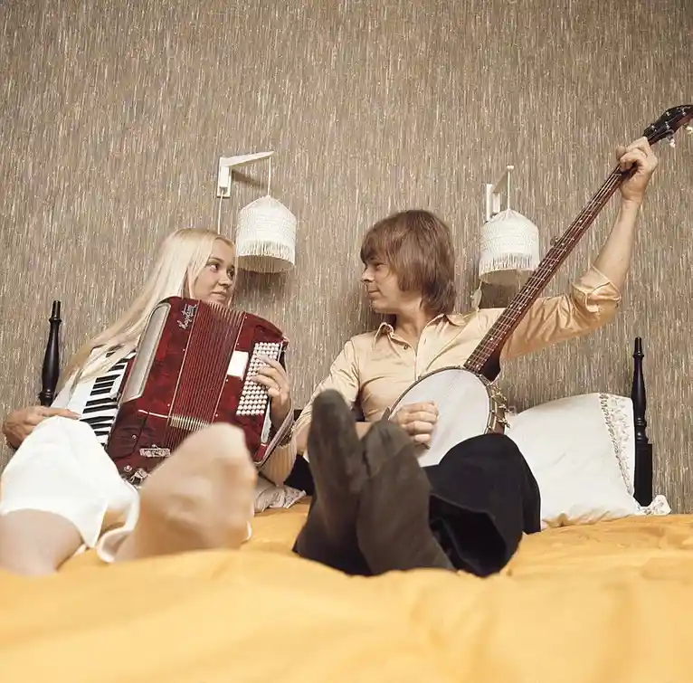 A vintage photo shows a man and woman sitting on a bed, each holding a musical instrument — the woman an accordion and the…