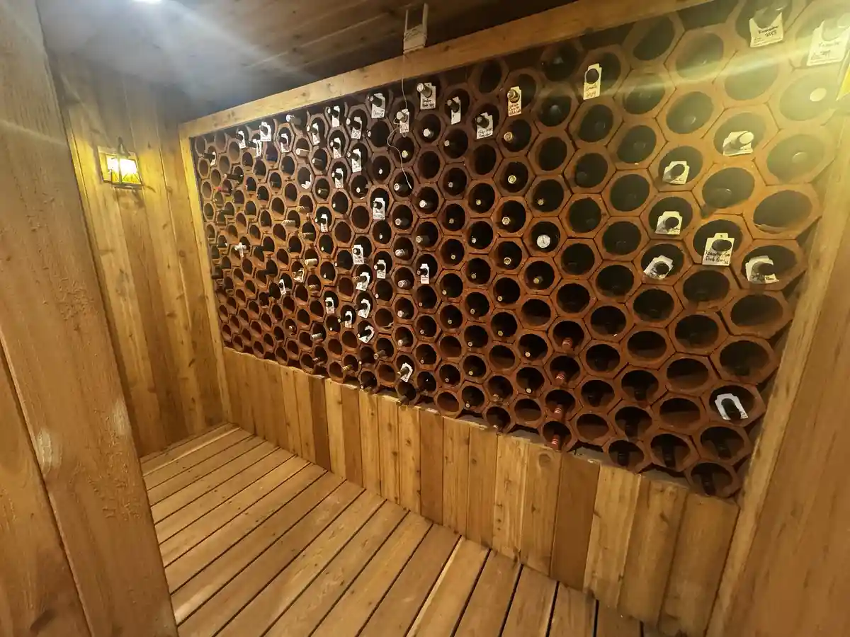 a wooden wine cellar room with a large wall of hexagonal slots holding wine bottles, some slots are empty, the room has…