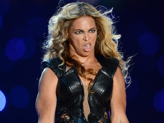 A woman with long blonde hair wearing a black leather-like outfit stands against a blue bokeh background, making a fierce…