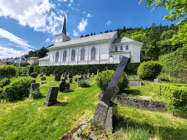 A bright, sunny photo of a white church with a steeple behind a well-kept cemetery filled with gravestones. One prominent…