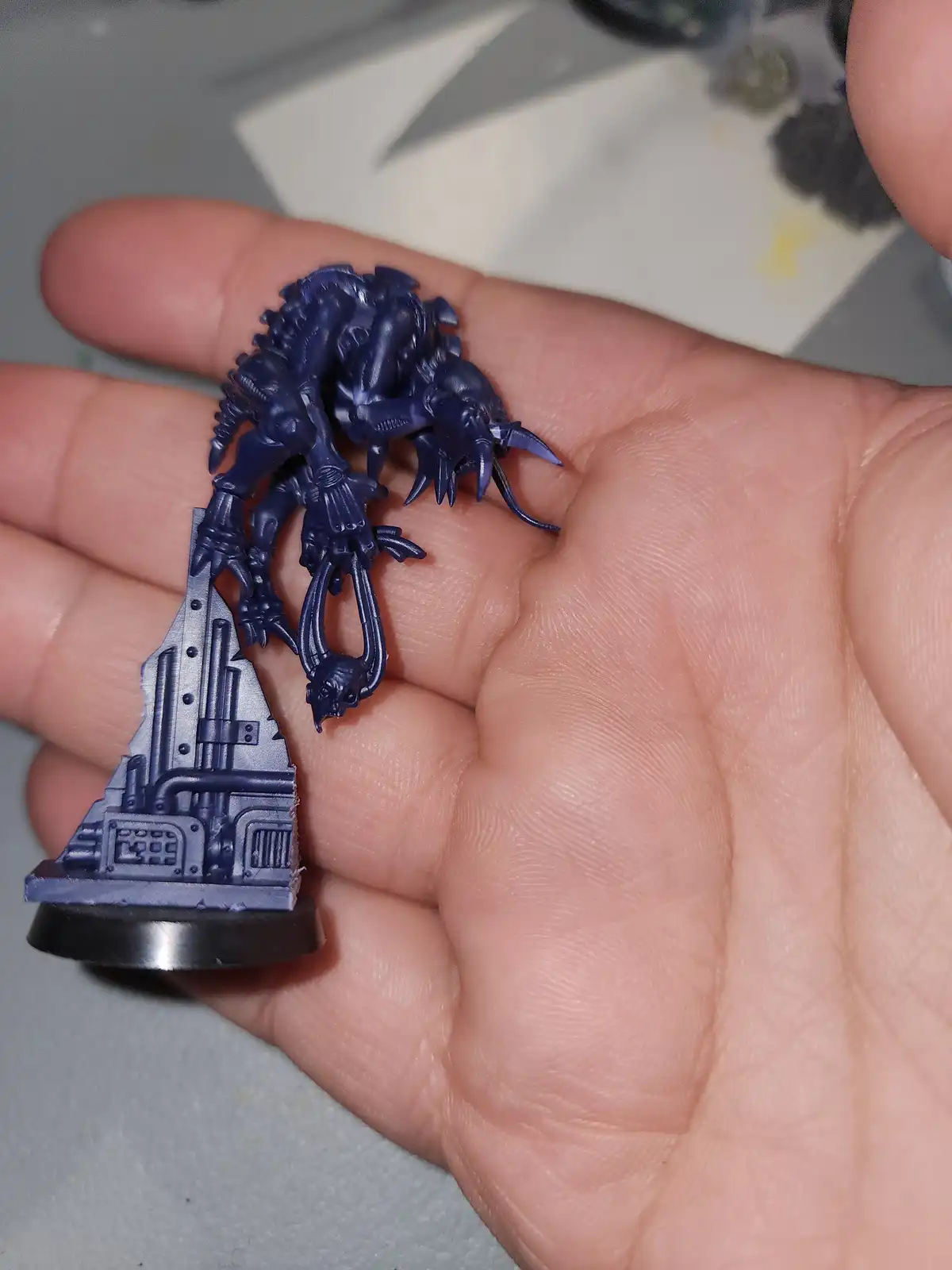 A person holds a small, unpainted miniature model of a monstrous creature with multiple limbs and claws, which has been…
