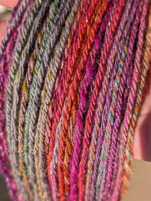 A close-up photo shows a hand holding a skein of multicolored, sparkly yarn with vibrant shades of pink, orange, purple, and…