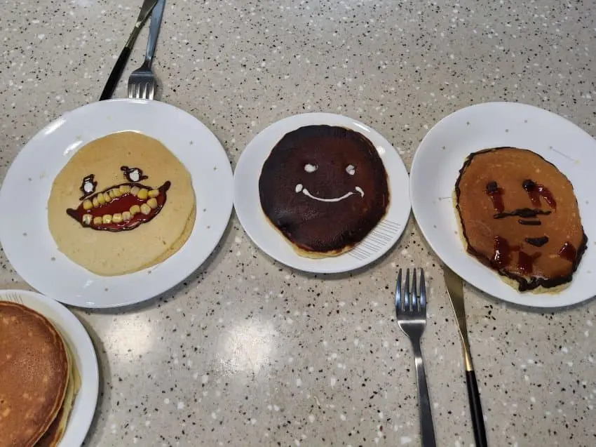 Three pancakes on white plates arranged on a speckled countertop, each decorated with syrup and butter to resemble faces…