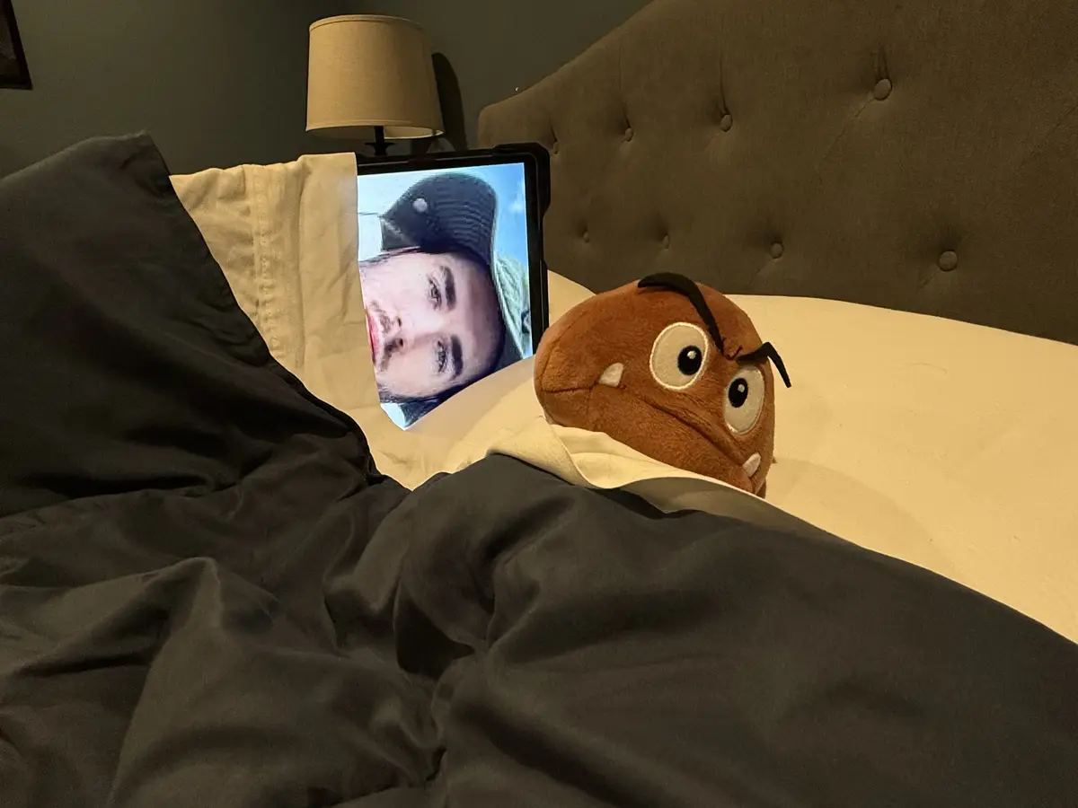 A brown plush toy with large eyes and angry eyebrows sits in bed under a dark blanket, next to a tablet displaying a…