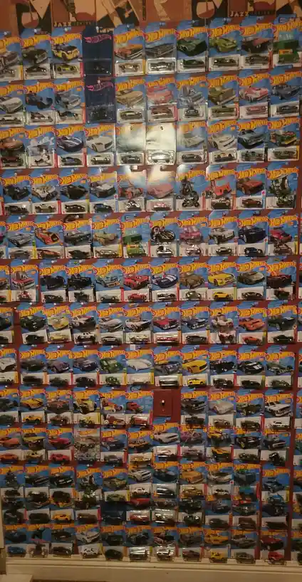 A wall is densely covered with dozens of Hot Wheels toy cars in their original blue and red packaging, arranged in a grid…