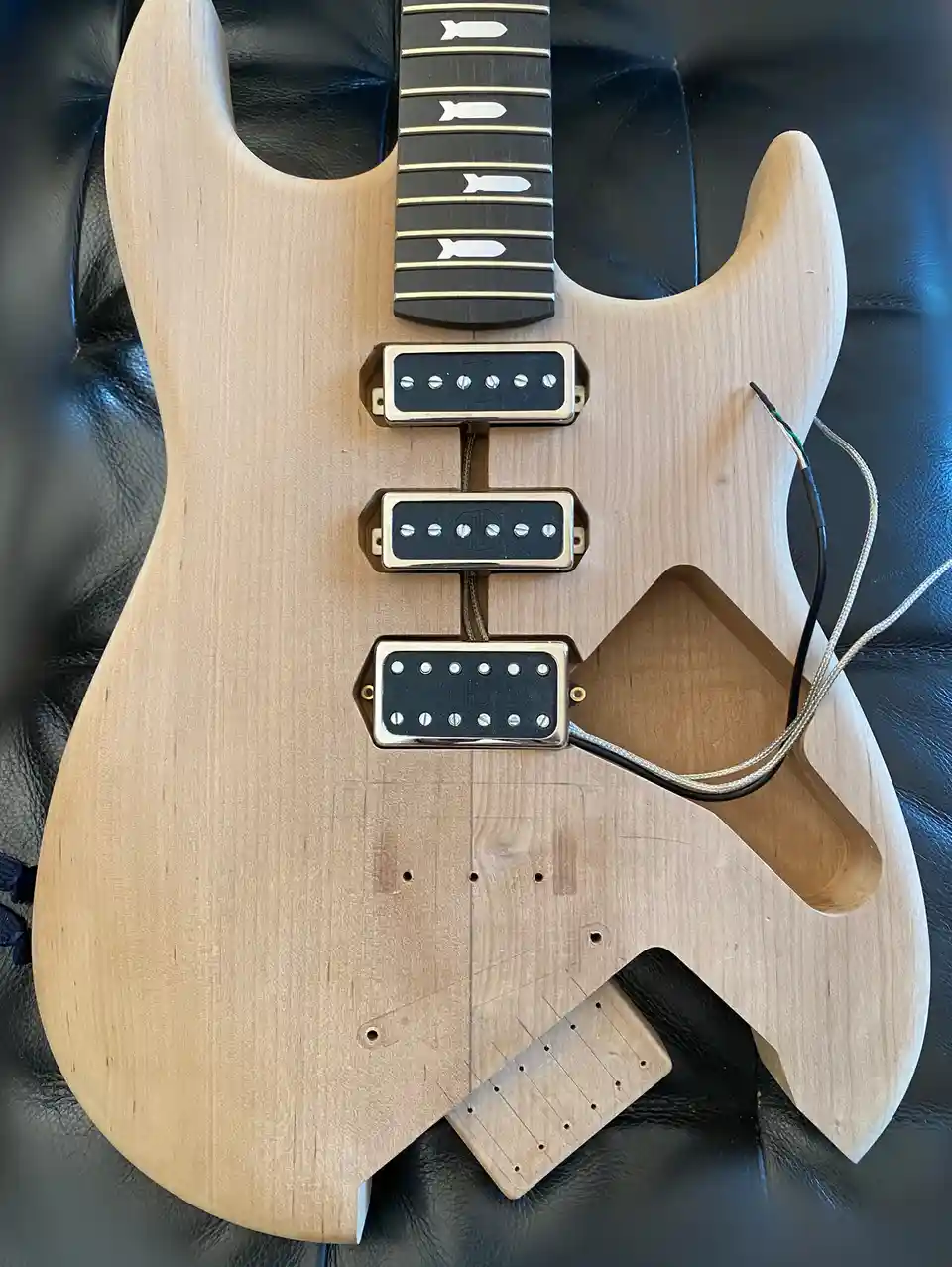 A light-wood, unfinished electric guitar body with three pickups and exposed wiring rests on a black leather surface. The…