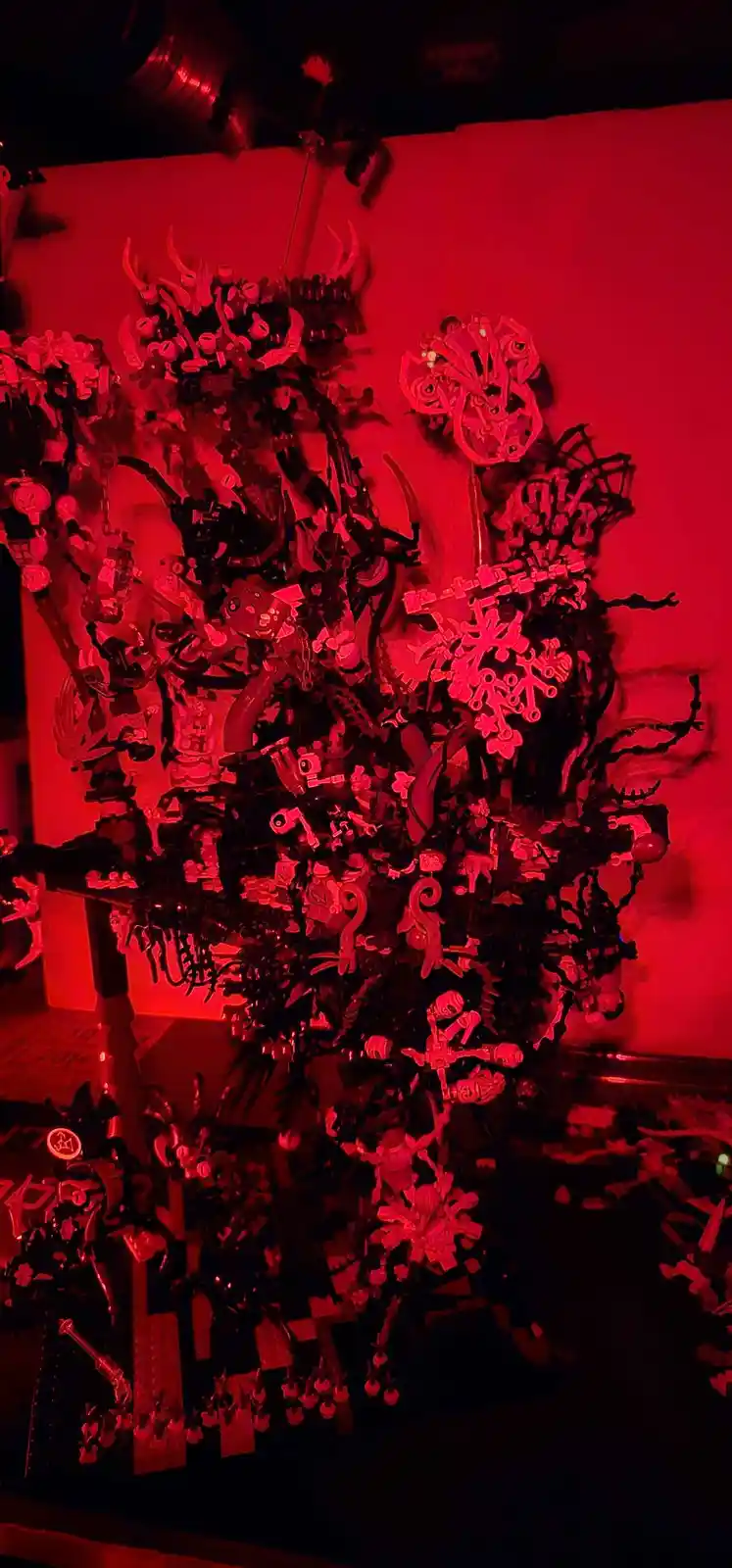 A Christmas tree made entirely of Lego pieces, illuminated by red lighting, creating a dark, demonic aesthetic. The tree is…