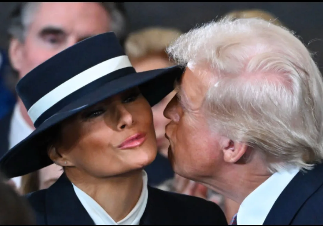A man with white hair kisses a woman wearing a black hat with a white band on the cheek. The woman has a neutral expression…