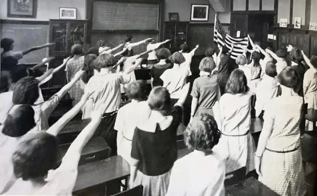 A black-and-white photo shows a classroom of students from the 1950s or 60s standing at their desks with their right arms…