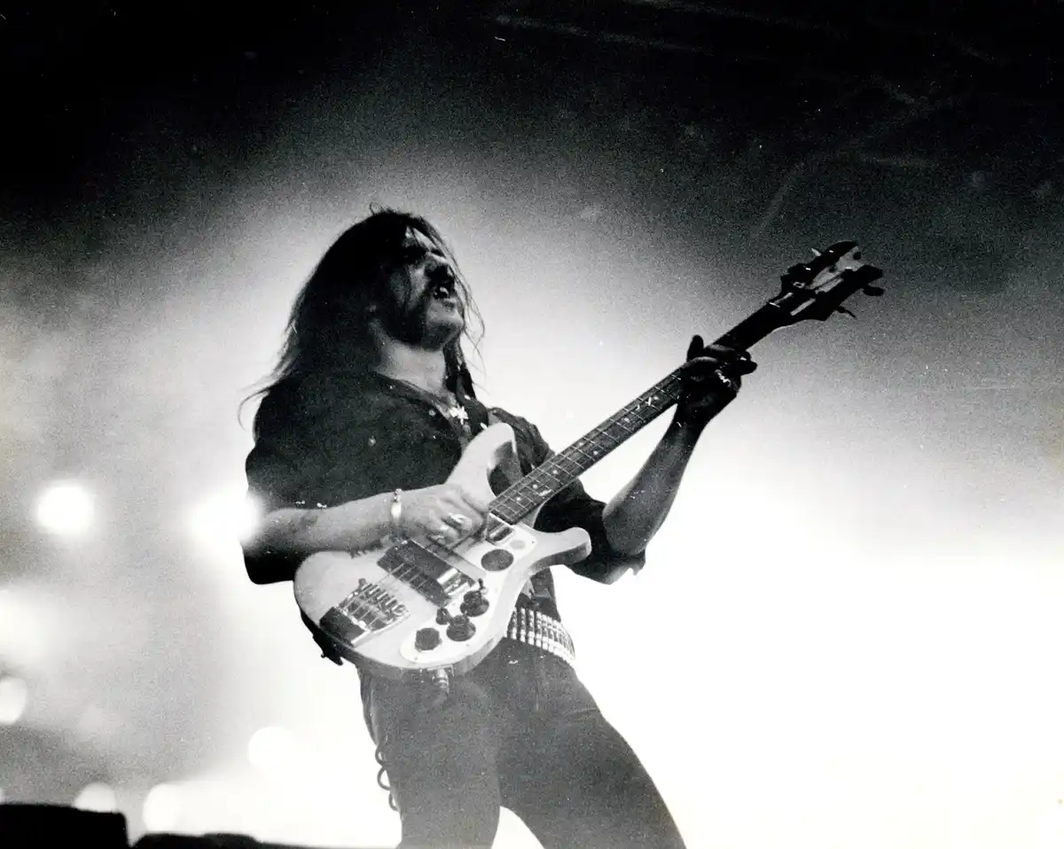 A black and white photo shows a long-haired man with a beard, intensely playing an electric guitar on stage under bright…