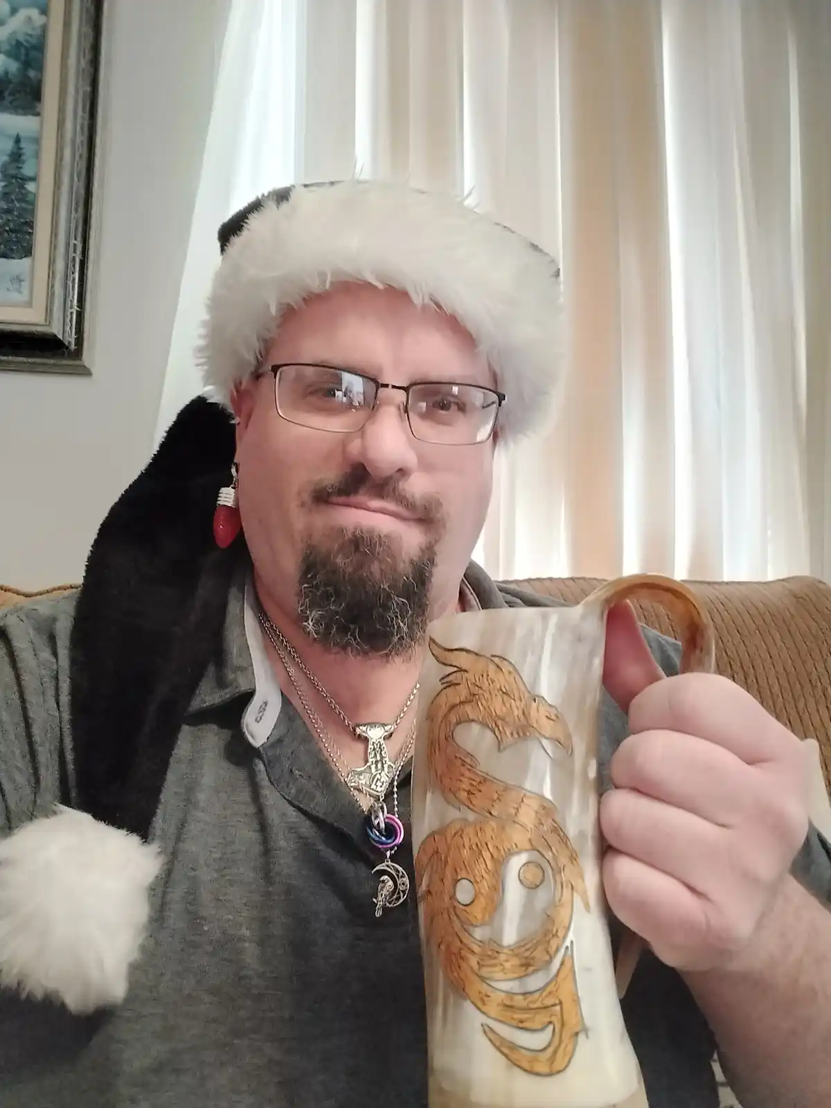 A man wearing a Santa hat and glasses holds up a mug with a dragon design, looking at the camera with a slight smirk. The…