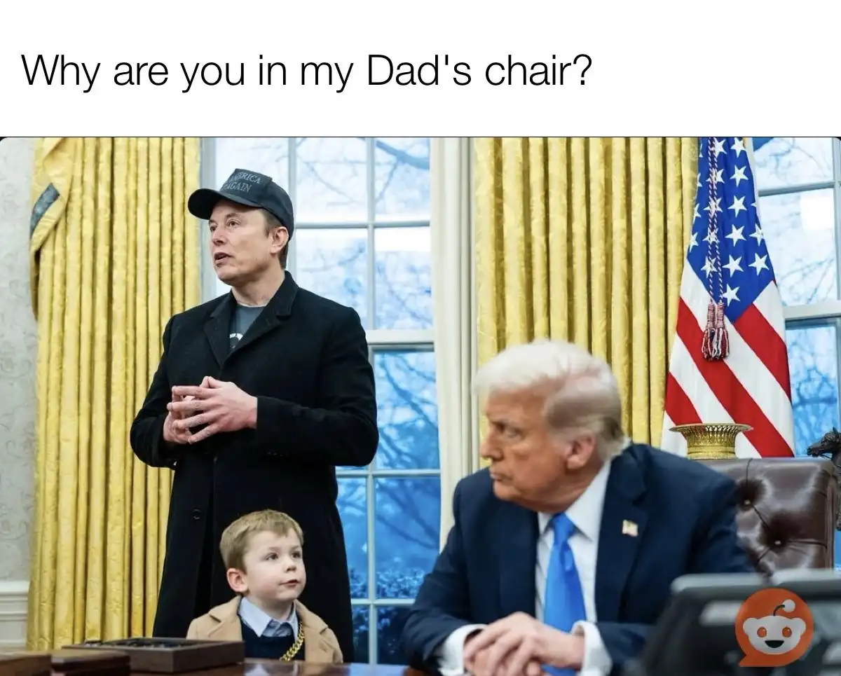 A young boy in a tan coat and gold chain sits at a desk in the Oval Office, looking up at Elon Musk who stands behind him…