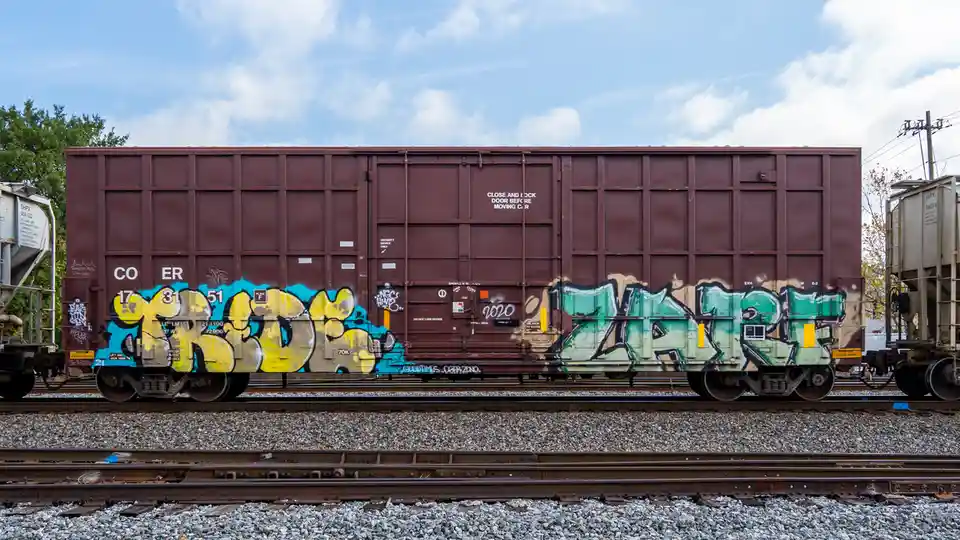 A maroon freight train car covered in colorful graffiti sits on railway tracks under a partly cloudy sky. The car has…