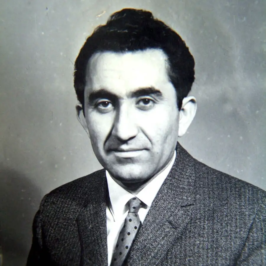 A black-and-white portrait of a man with dark hair, wearing a suit and tie, looking directly at the camera with a neutral…