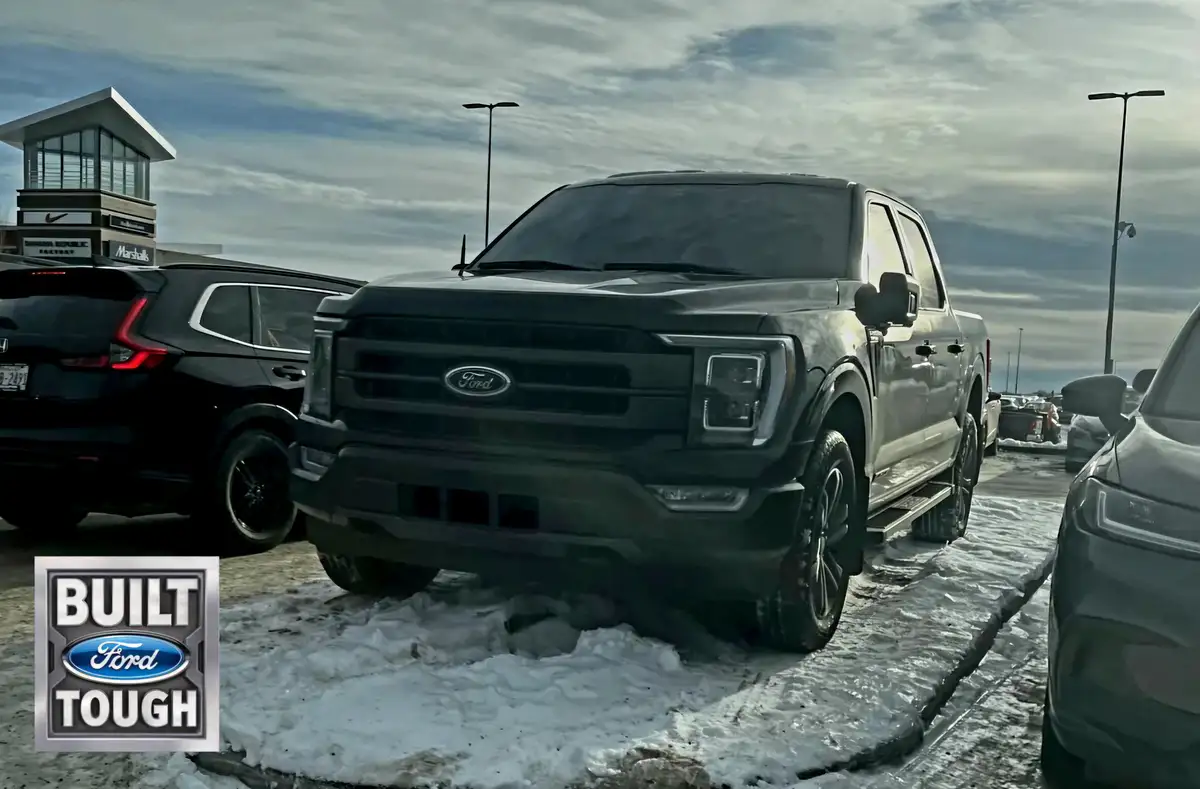 A black Ford F-150 pickup truck is parked in a snowy lot, partially stuck in a snowbank, with other cars nearby and a…