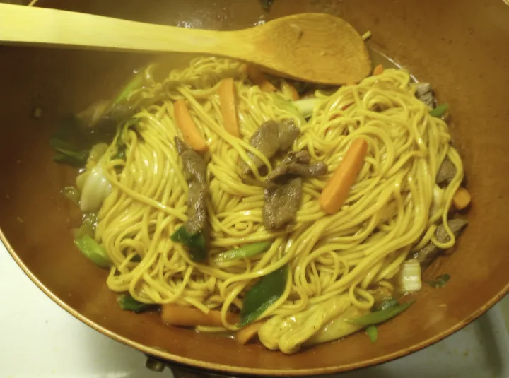 A close-up photo of stir-fried noodles in a wok with vegetables and meat, a wooden spoon resting on top. Overlaid text…