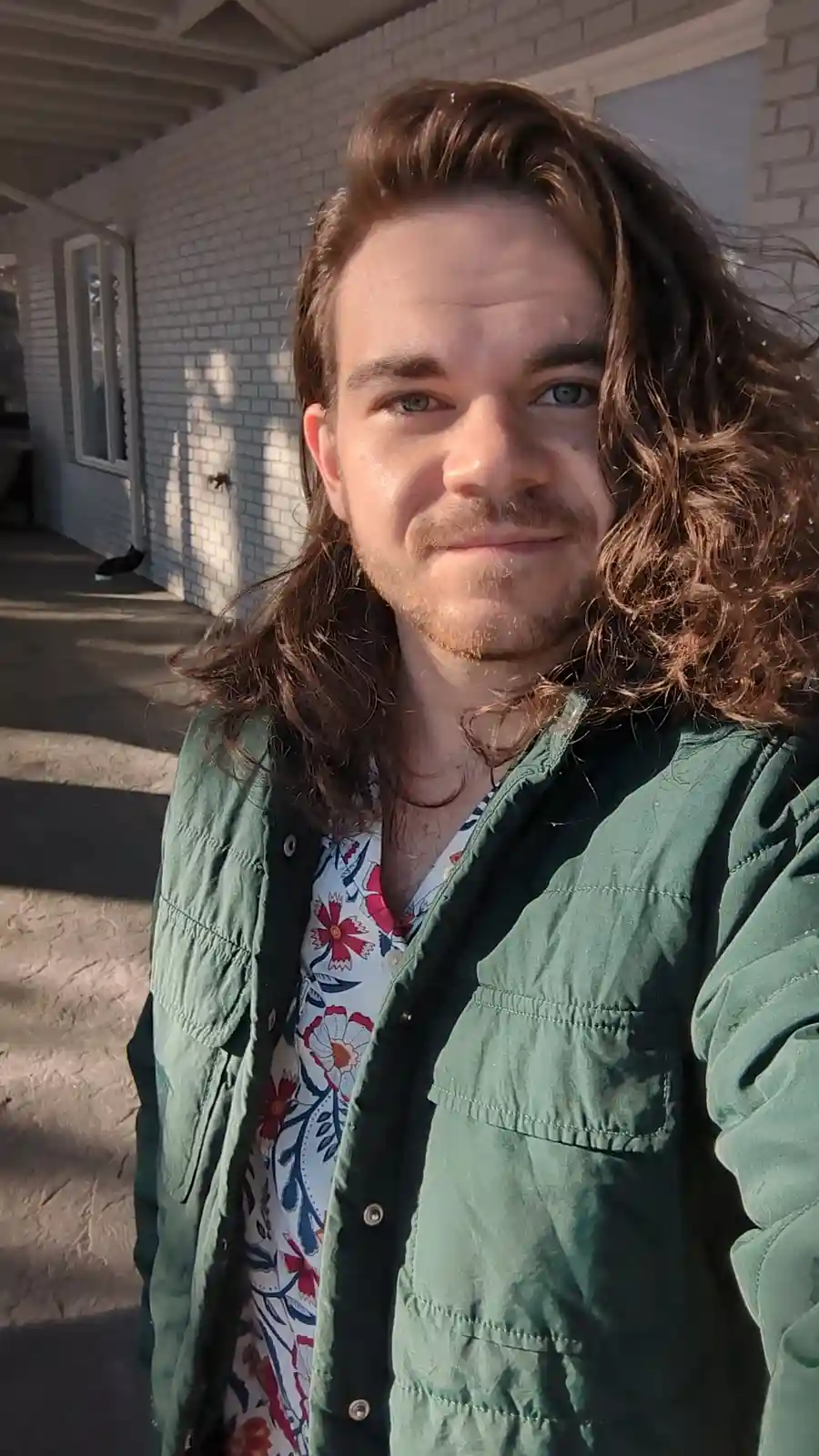 A man with long brown hair and a mustache stands outside a white brick house, wearing a green jacket over a floral shirt…