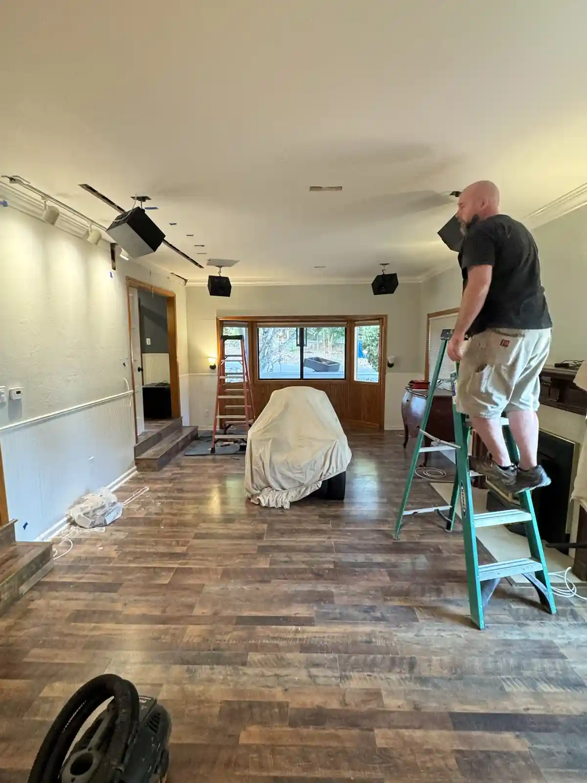 A bald man in a black t-shirt and khaki shorts stands on a green stepladder inside a room with hardwood floors, installing…