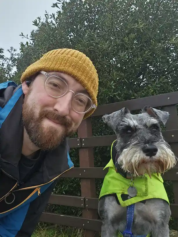 A man with a beard, yellow beanie, and clear glasses smiles at the camera while sitting on a wooden bench next to a gray and…
