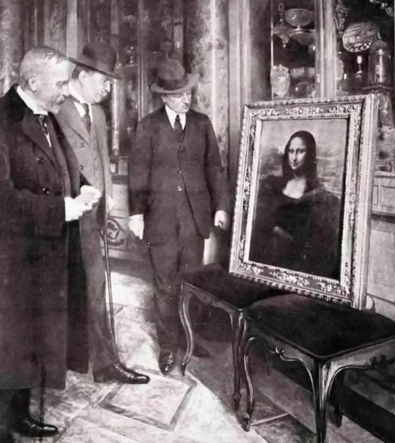 A black-and-white historical photograph shows three men in early 20th-century formal attire standing in an ornate room…