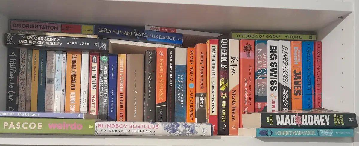 A close-up photo of a white bookshelf filled with numerous books, their spines showing varied colors and titles like 'Mad…