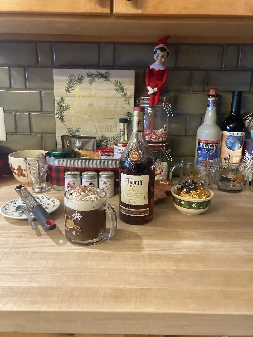 A kitchen counter is decorated for Christmas with a mug of hot chocolate topped with whipped cream, a bottle of Asbach Uralt…
