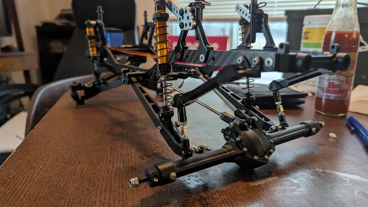 A close-up photo of a black RC car chassis with gold coil springs sitting on a wooden table, next to a bottle of 'Original…