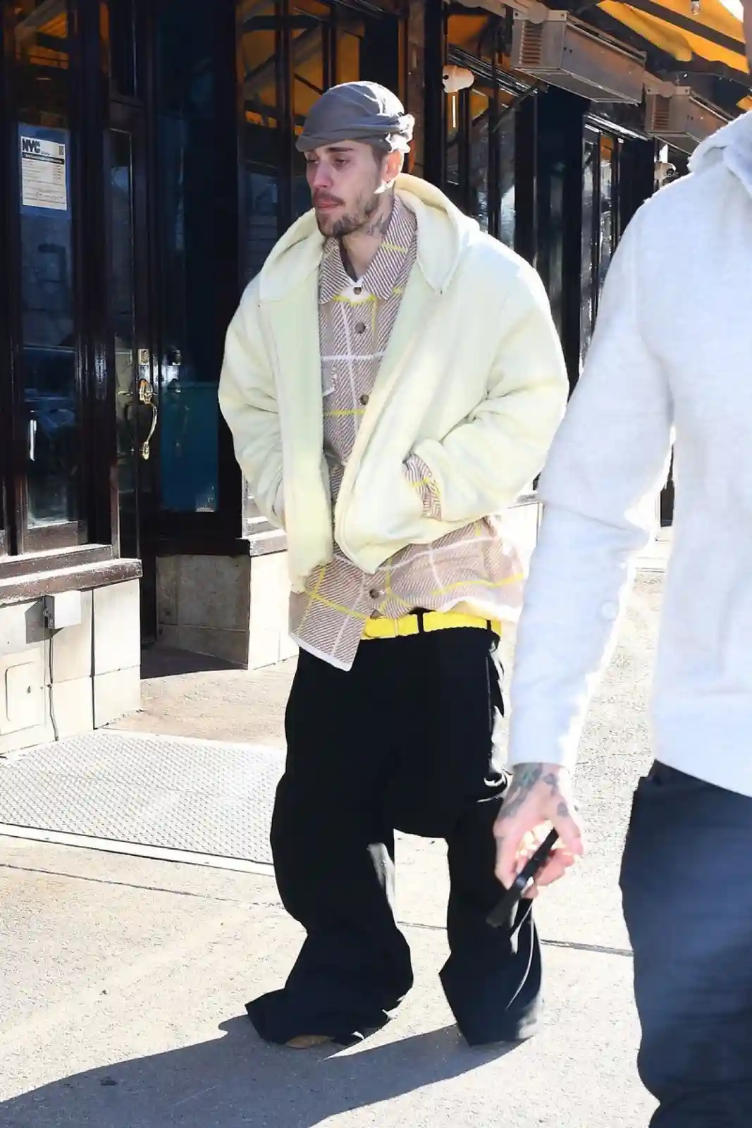 A man wearing a grey cap, a pale yellow fleece jacket over a plaid shirt with a yellow belt and black baggy pants, stands…