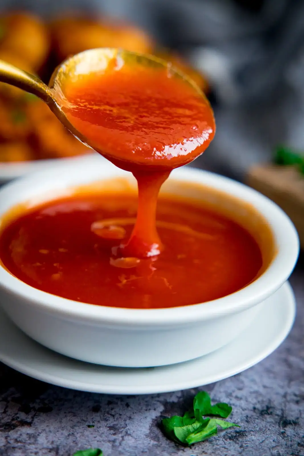 A close-up photo shows a golden spoon lifting thick, glossy red sauce from a white ceramic bowl, with the sauce dripping…
